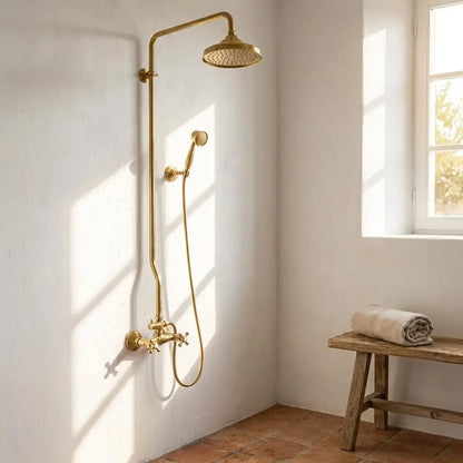 Unlacquered brass exposed pipe shower system with rainfall head and handheld sprayer mounted on a textured white plaster wall next to a wooden bench.