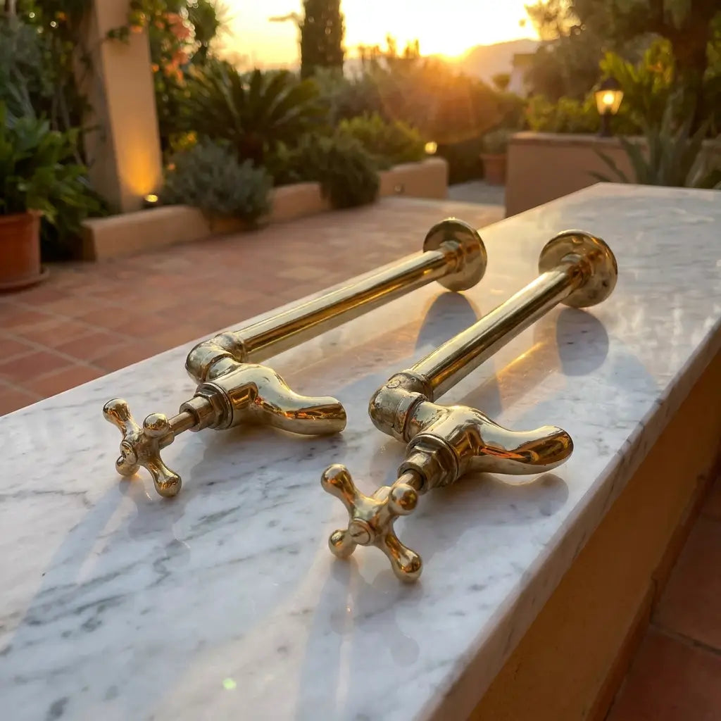 Two unlacquered brass water dispenser faucets resting horizontally on a white marble ledge outdoors at sunset