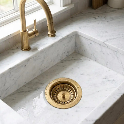 Unlacquered brass kitchen sink strainer and standard 3.5 inch solid brass drain basket installed in a white marble countertop sink.