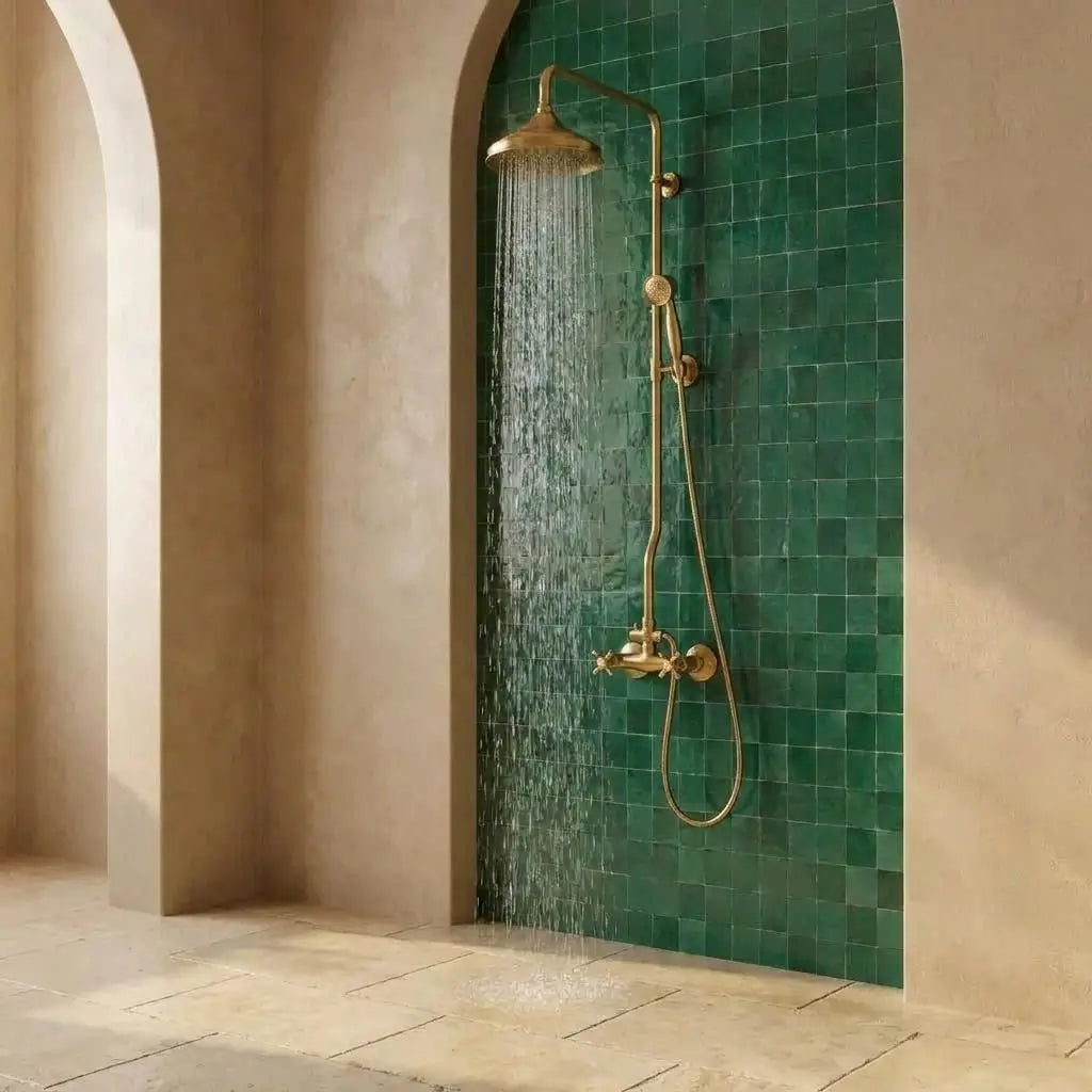Unlacquered brass exposed pipe shower system with flowing rainfall water, mounted on a green zellige tile wall inside a luxury arched bathroom alcove.