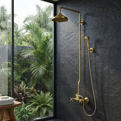 Unlacquered brass exposed pipe shower system with a rainfall head and handheld sprayer mounted on a dark textured slate wall next to a large window overlooking tropical plants.