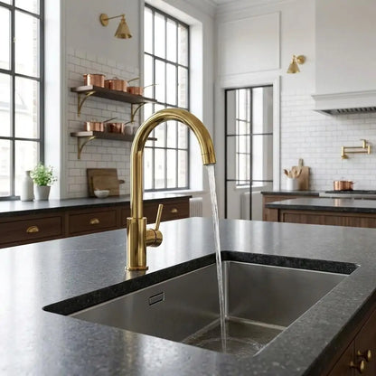 Unlacquered brass curved sink faucet with running water flow installed on a kitchen island.