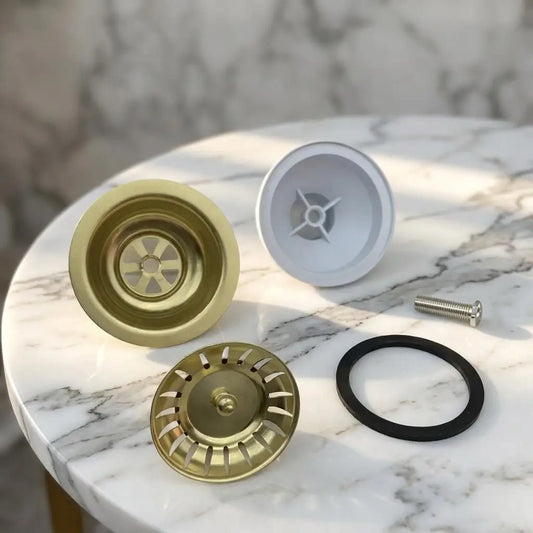 Complete components of an unlacquered brass kitchen sink waste kit including the main flange, removable strainer basket, rubber sealing ring, and screw on a marble surface.