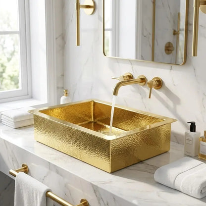 A rectangular vessel brass Moroccan sink sitting fully on a white marble countertop with running water from a modern wall-mounted gold faucet.