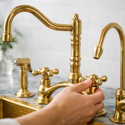 A person's hand turning the cross handle of an unlacquered brass Victorian classic kitchen bridge faucet set, featuring a side sprayer and a cold water filter tap.