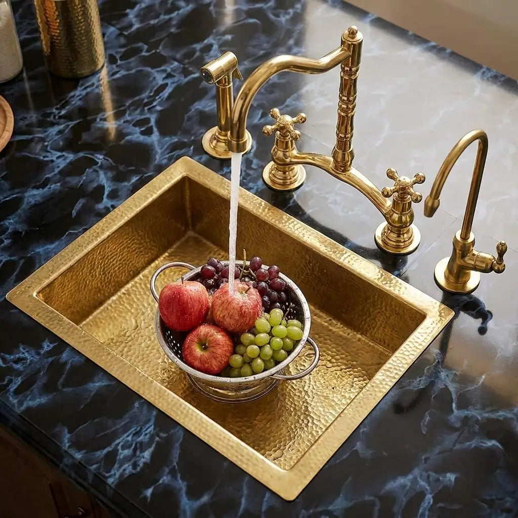 Unlacquered brass Victorian classic kitchen bridge faucet with running water into a gold sink filled with a basket of fresh apples and grapes on a dark marble countertop.