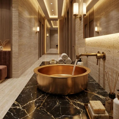 Water flowing from a wall-mounted brass faucet into a handcrafted round brass sink on a black marble countertop in a luxury hotel hallway with textured stone walls.